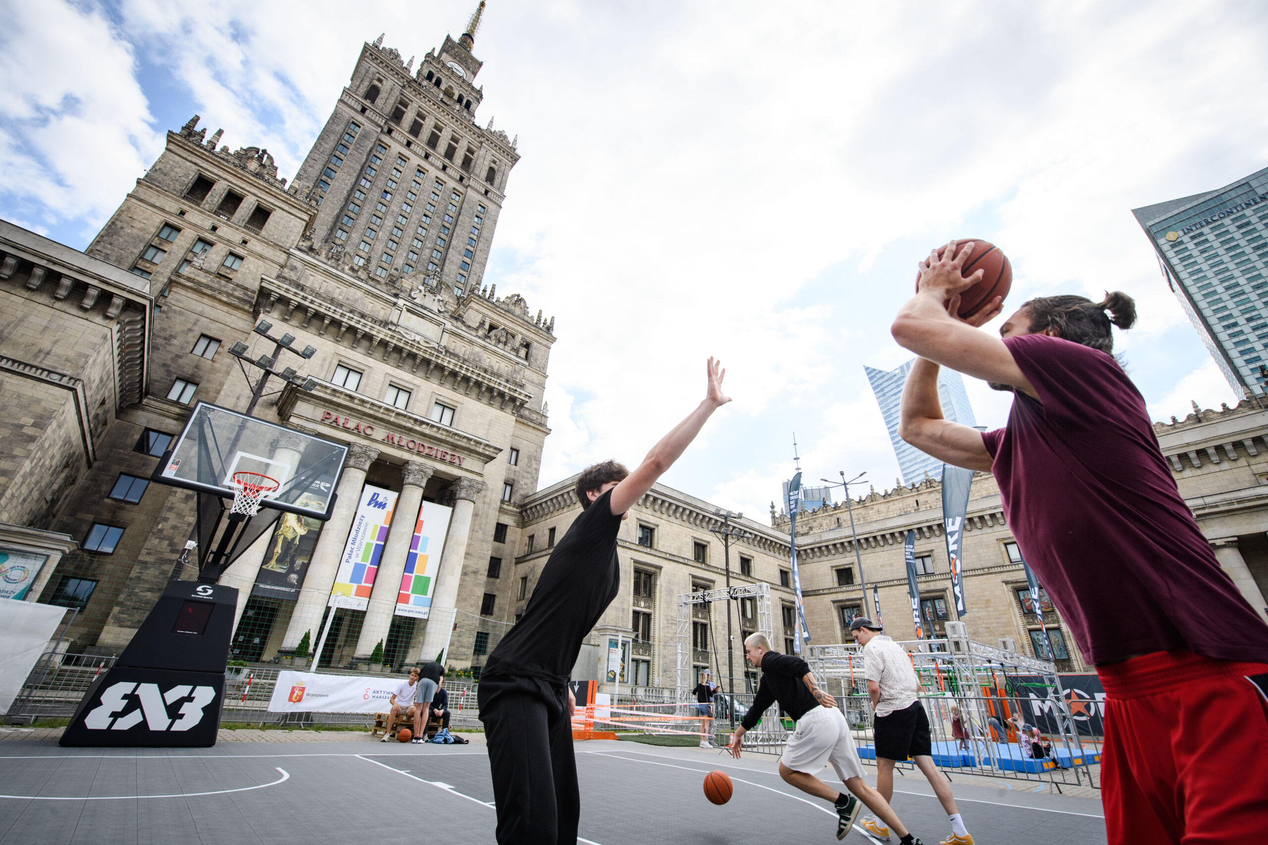 Na pierwszym planie kilka osób gra w koszykówkę 3x3 na boisku ustawionym przed Pałacem Kultury i Nauki w Warszawie. Jeden z uczestników oddaje rzut do kosza, a piłka znajduje się w powietrzu. W tle widoczna jest charakterystyczna bryła Pałacu Kultury i Nauki, boisko z koszem do gry oraz elementy infrastruktury Rodzinnej Strefy Sportu. Zdjęcie wykonano z niskiej perspektywy, co podkreśla wysokość budynku i dynamikę rozgrywki. Niebo jest częściowo zachmurzone.