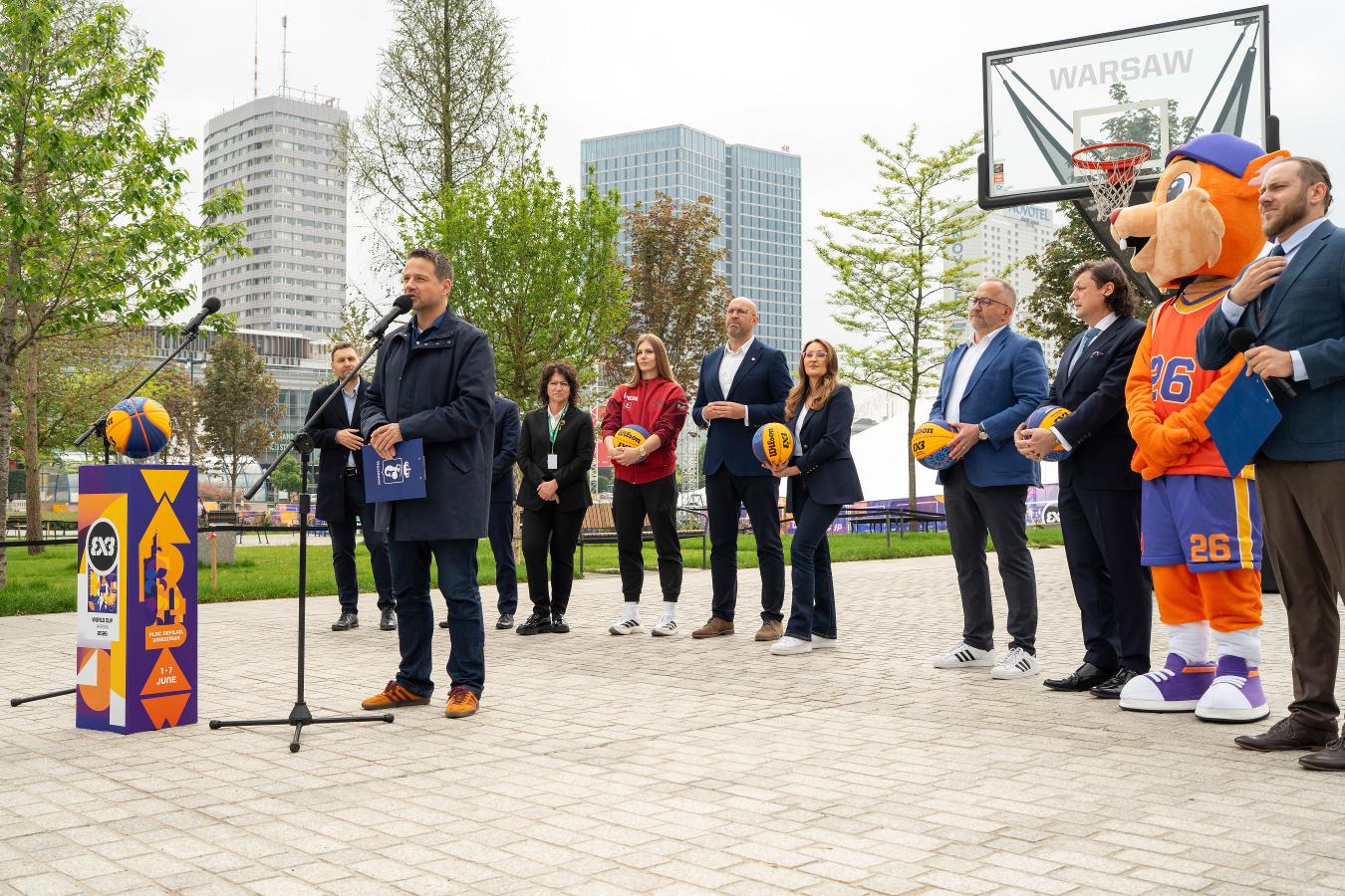 Oficjalna konferencja prasowa dotycząca Mistrzostw Świata FIBA 3x3 przy boisku do koszykówki na Placu Defilad w Warszawie. Na pierwszym planie przemawia mężczyzna stojący przy mikrofonie, wokół niego znajdują się przedstawiciele organizatorów i partnerów wydarzenia, część trzyma piłki do koszykówki 3x3. W tle widoczne są kosz do gry, miejska zabudowa centrum Warszawy oraz maskotka wydarzenia.