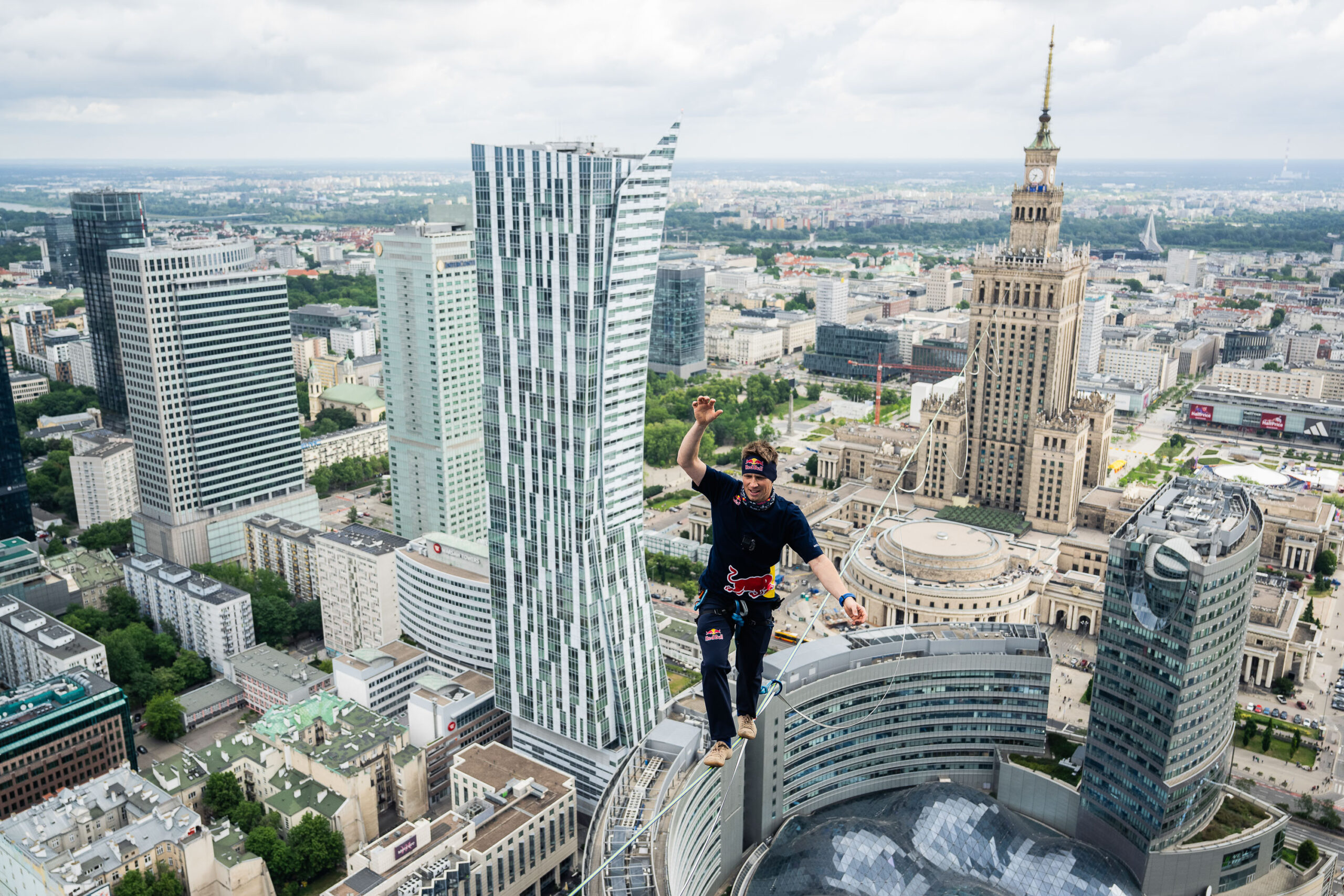 Slackliner przechodzi po linie rozpiętej pomiędzy warszawskimi wieżowcami, kilkaset metrów nad ziemią. W tle rozciąga się panorama Warszawy z dominującym Pałacem Kultury i Nauki oraz nowoczesną zabudową centrum miasta. Zdjęcie podkreśla wysokość, na jakiej odbywa się przejście, oraz rozległy widok na stolicę.